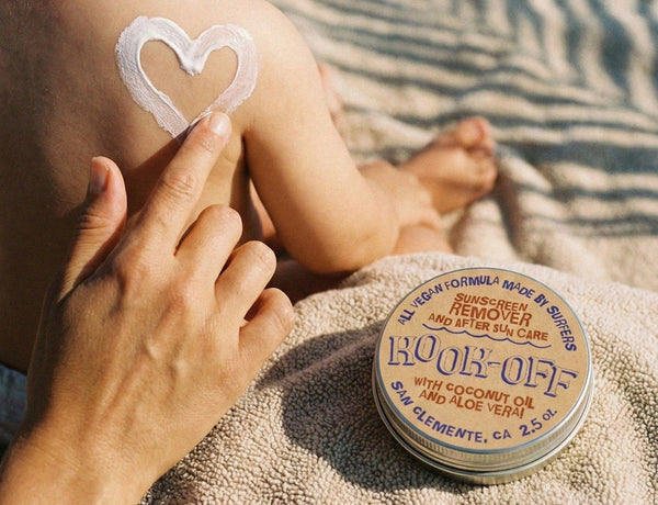 Hand drawing a sunscreen heart on a baby's back at the beach next to a tin of Kook-Off sunscreen remover