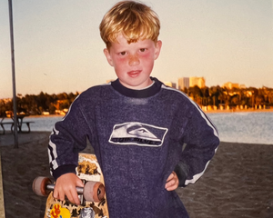 Young boy with sunburned cheeks and nose, holding a skateboard at the beach during sunset, wearing a Quiksilver sweatshirt.