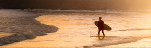 Surfer walking into the ocean at golden hour, carrying a surfboard and silhouetted against the glowing sunset.