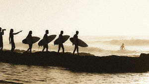 Silhouettes of surfers walking along a rocky shoreline at sunrise, heading toward the ocean as one surfer waits in the water.